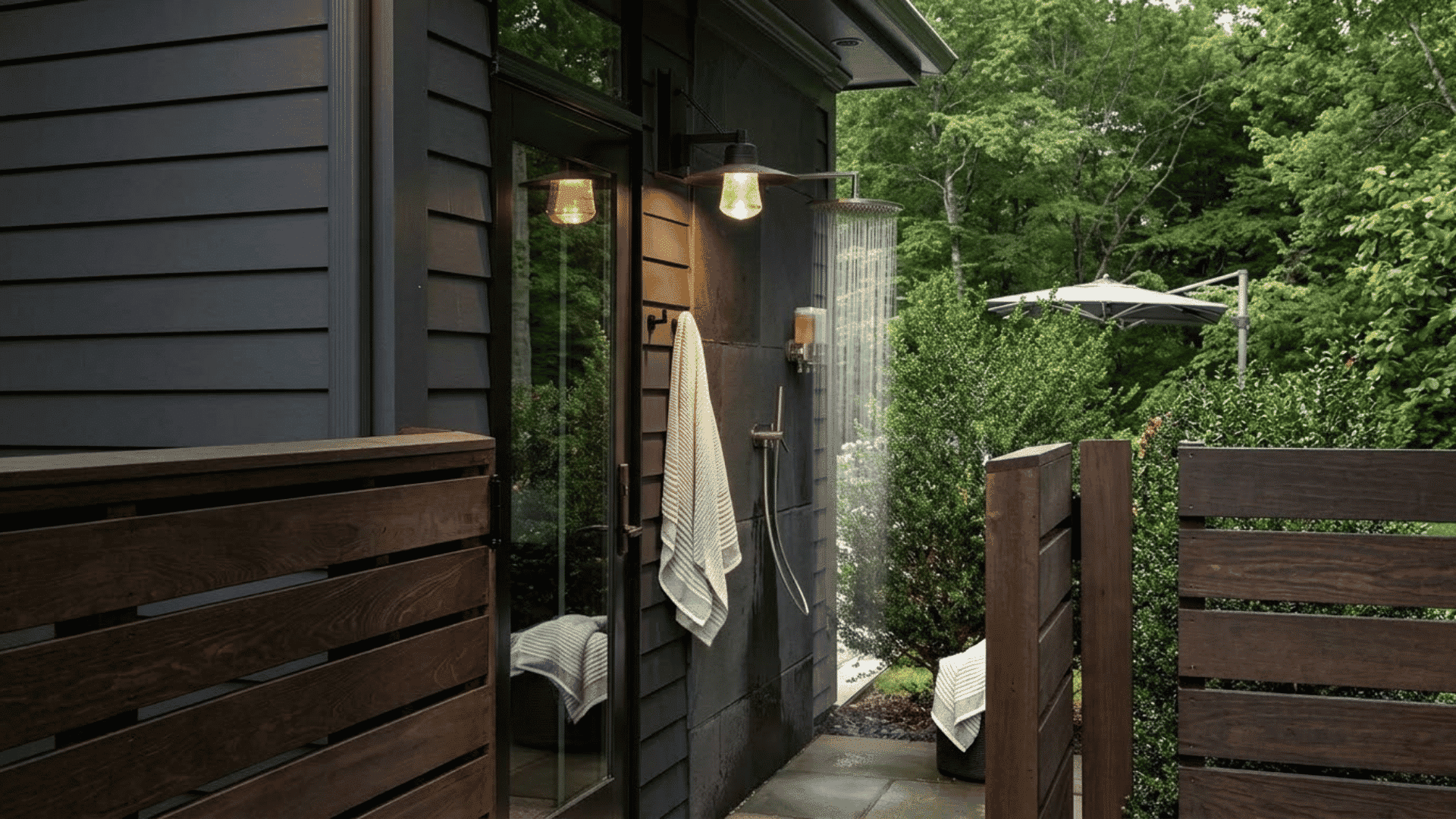 Modern outdoor shower attached to a dark exterior wall with wooden privacy panels, towel, glass door, and garden surroundings.