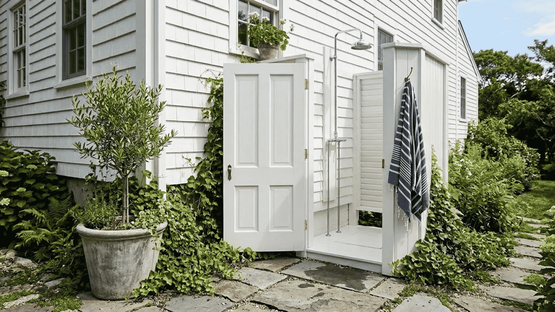 white outdoor shower stall beside a house with privacy panels, striped towel, stone pathway, potted plant, and garden greenery